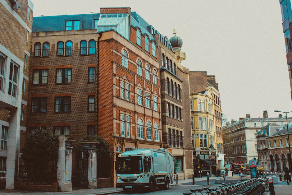 A light blue delivery van parked on a city street with multi-storey buildings in the background, including a red brick building with arched windows and a building with cream-colored facades and bay wi